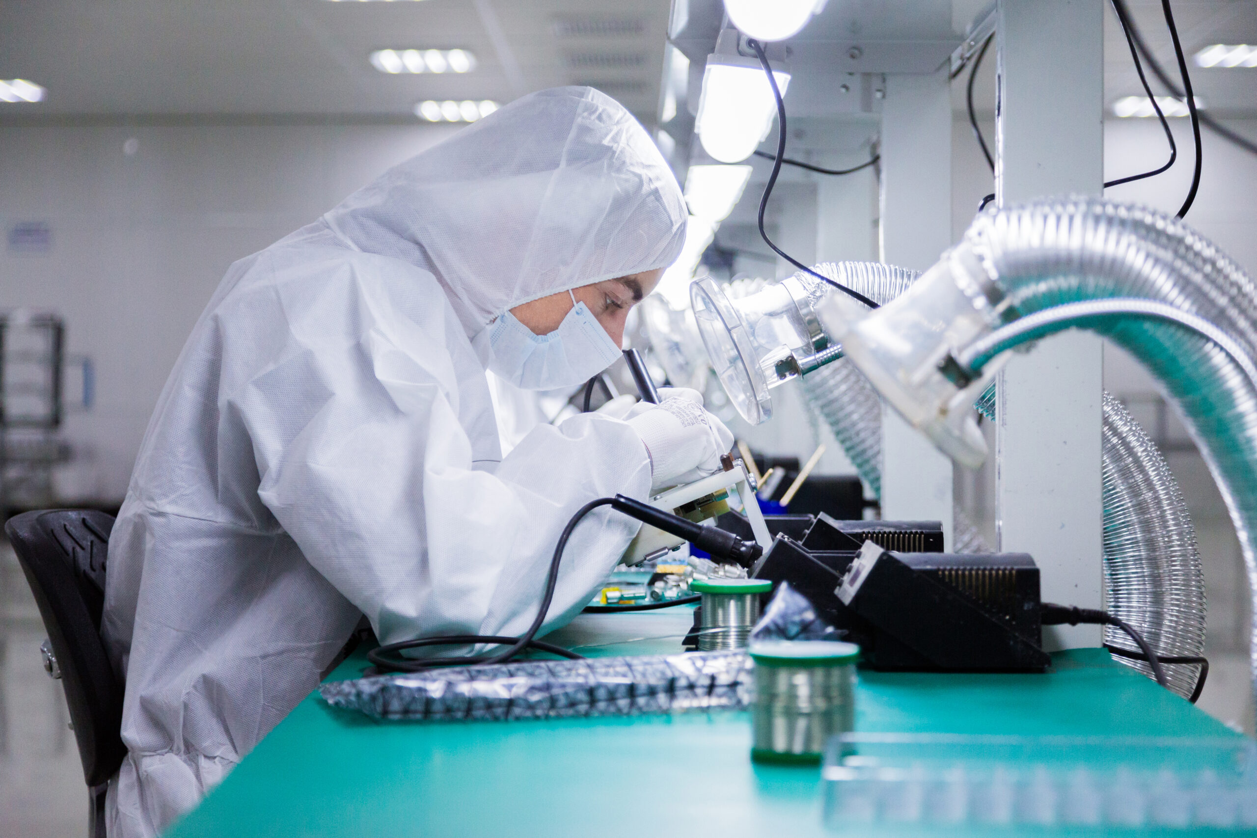 factory workers in white lab suits and face masks, sitting on the chairs are producing tv sets with soldering irons under bright lamps