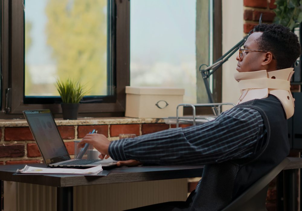 Business man wearing cervical neck collar at work after vertebrae injury in startup office. Injured employee using medical foam while he works with laptop to plan commercial investment.