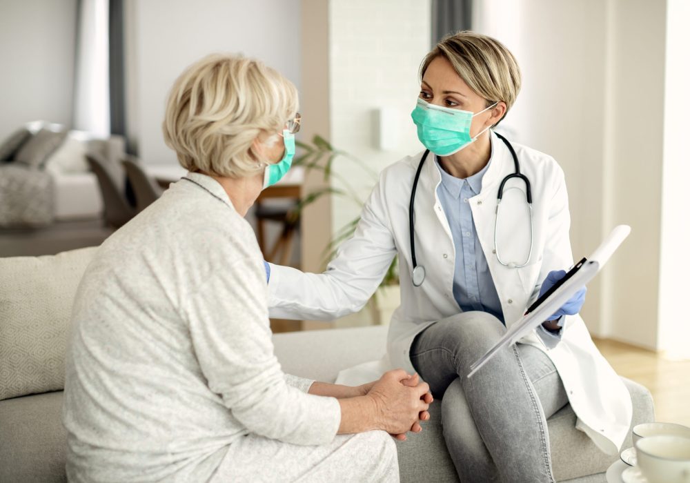 Female doctor and senior woman wearing protective face masks while communicating during home visit.