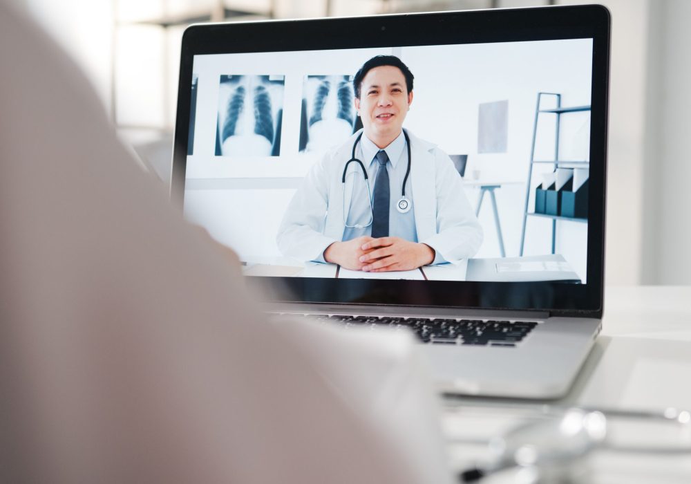 Young Asia male doctor in white medical uniform using laptop talking video conference call with senior doctor at desk in health clinic or hospital. Social distancing, quarantine for corona virus.